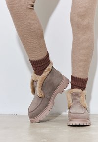 Bottines en suède gris avec un col en fourrure beige et une semelle en caoutchouc rose, assorties à des chaussettes marron côtelées. Texture lisse et design décontracté.