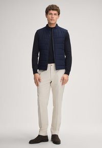 Granatowa pikowana kamizelka z wysokim kołnierzem, zestawiona z czarną bluzką z długim rękawem i jasnobeżowymi spodniami. Brązowe zamszowe botki do kostki dopełniają stylizację.