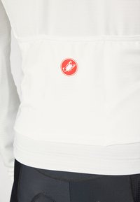 Nahaufnahme einer Person, die eine weiße strukturierte Jacke mit einem roten kreisförmigen Krabbenlogo im unteren Rückenbereich trägt, kombiniert mit schwarzen Hosen.