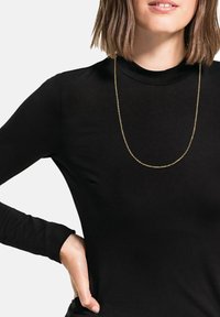 Collana in oro appesa sopra una maglia nera a maniche lunghe, con un design aderente e una superficie liscia su uno sfondo neutro.