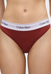 Μπορντό μπικίνι Calvin Klein με λευκή μέση που φέρει μαύρο λογότυπο, φορεμένο από άτομο που δείχνει την κοιλιά και το πάνω μέρος των μηρών.