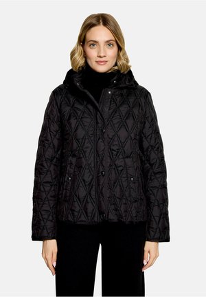 Femme portant une veste matelassée noire avec un col haut et des boutons sur le devant, debout devant un fond clair uni.