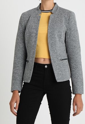 Veste ajustée grise avec un col haut, texture subtile et deux poches zippées, portée par-dessus un crop top côtelé jaune et un pantalon noir.