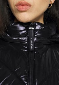 Gros plan sur une personne portant une doudoune Calvin Klein noire brillante avec une fermeture éclair, une boucle d'oreille en or en forme d'anneau visible.
