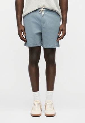 Man wearing light blue drawstring shorts, white crew socks, and beige sneakers, standing against a plain white background.