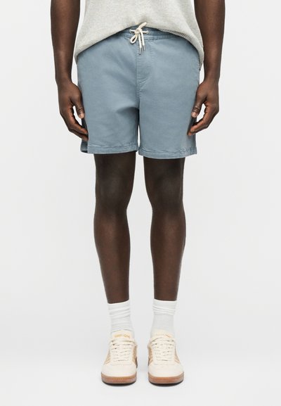 Homme portant un short à cordon de serrage bleu clair, des chaussettes blanches montantes et des baskets beiges, debout devant un fond blanc uni.