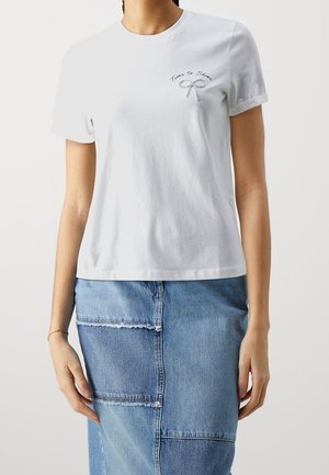 Femme portant un t-shirt blanc avec le texte "Time to Shine" et un nœud en strass sur la poitrine, associé à une jupe en jean patchwork.