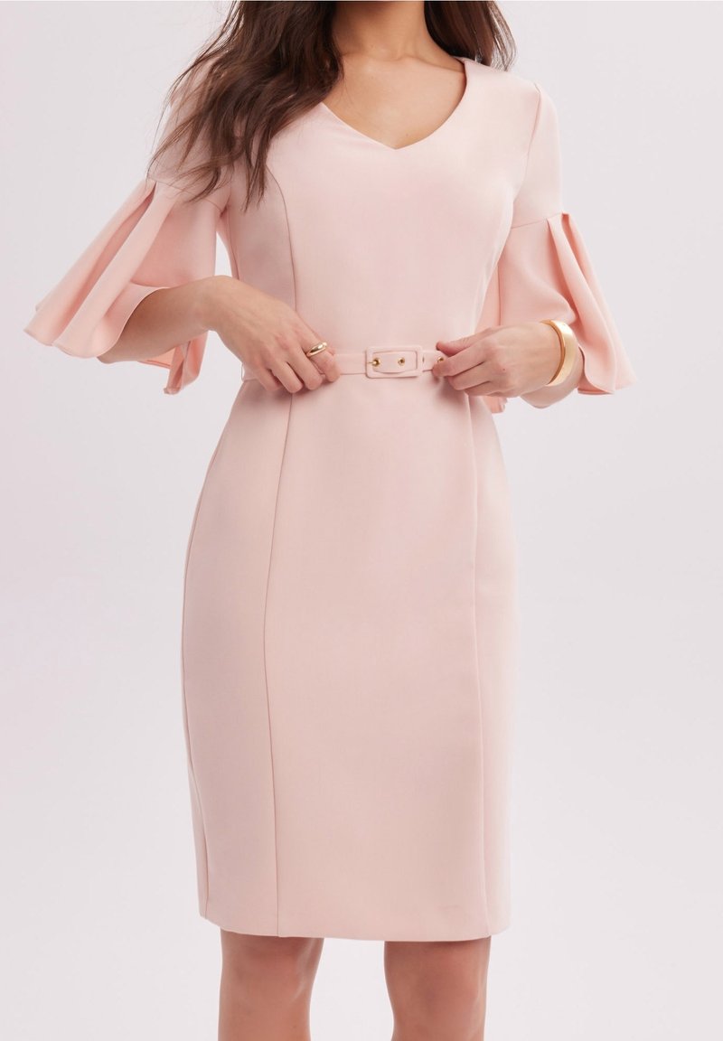 Femme ajustant sa ceinture sur une robe rose clair mi-longue avec un col en V et des manches évasées jusqu'au coude, portant un bracelet et une bague en or.