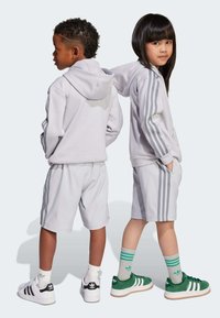 Ljusgrå huvtröja och shorts med tre vertikala ränder. Barnet har på sig gröna skor med vita detaljer och randiga strumpor.