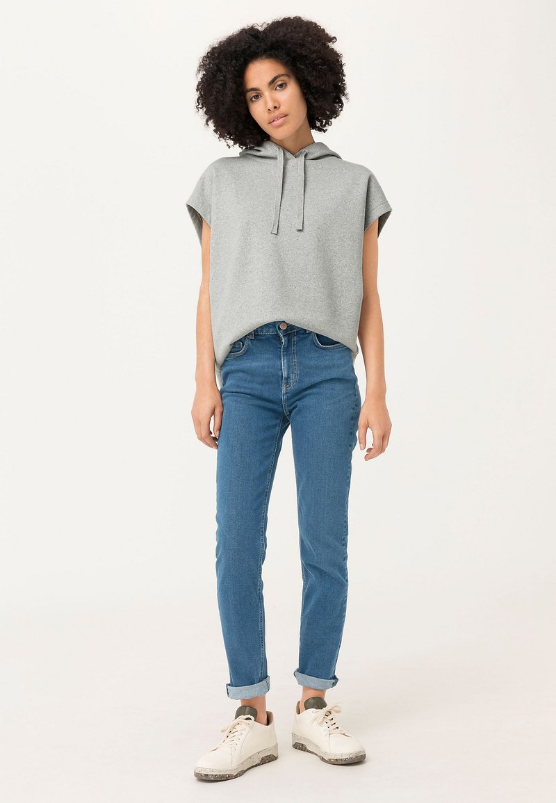 Grijze hoodie zonder mouwen met een relaxte pasvorm, gecombineerd met blauwe jeans met opgerolde manchetten en witte sneakers met grijze accenten.