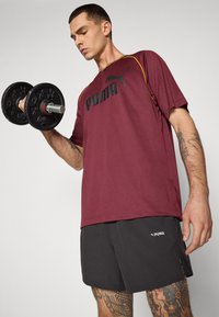Bordeaux sportt-shirt met zwart logo, gele accenten en een losse fit, gecombineerd met zwarte shorts. Model houdt een zwarte dumbbell vast.