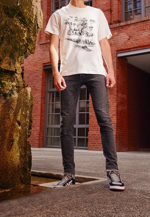 Person trägt weißes T-Shirt mit schwarzer Wildtierillustration, dunkle Skinny-Jeans und schwarz-weiße Sneakers und steht nahe eines moosbedeckten Felsens und eines Backsteingebäudes.