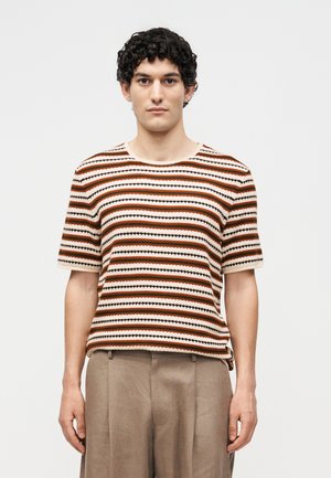 Ung mand med krøllet hår iført kortærmet striktrøje med brune, orange og sorte vandrette striber, kombineret med beige bukser.