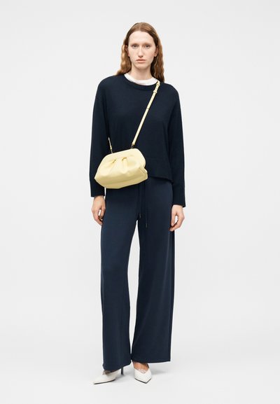 Femme portant un pull bleu marine et un pantalon à jambes larges avec des talons blancs, tenant un sac bandoulière jaune pâle sur un fond uni.