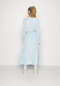 Vestido de manga longa azul claro com padrão floral, saia em camadas e cintura elástica. Apresenta um decote redondo e um corte fluido, combinado com botas brancas.