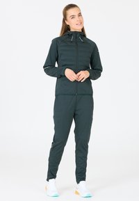 Donkergroen sportoutfit met een lichte, ritsbare jas met een gewatteerde bovenkant, bijpassende taps toelopende broek en witte sneakers met kleurrijke accenten.