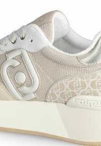 Sneaker con tessuto testurizzato beige, dettagli bianchi e logo. Presenta una suola spessa e un collare imbottito per il comfort.