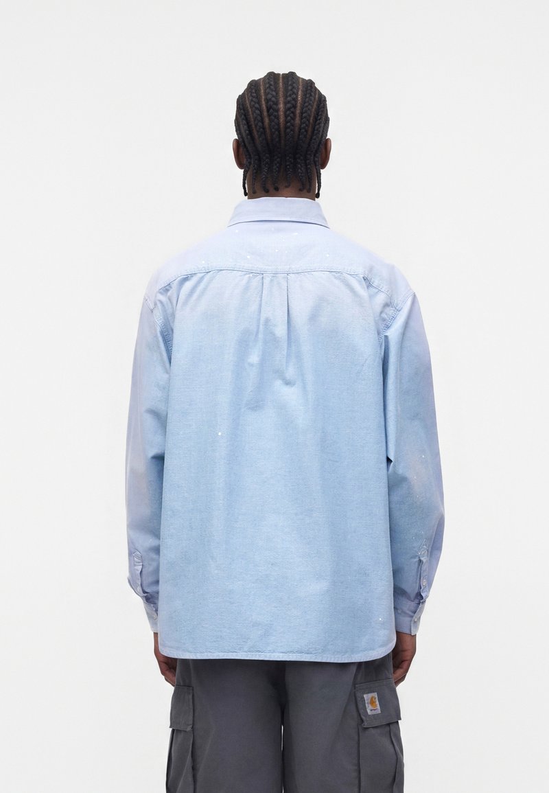 Uomo con capelli intrecciati che indossa una camicia azzurro chiaro a maniche lunghe e pantaloni cargo grigio scuro, in piedi di spalle su uno sfondo semplice.