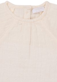 Licht beige kortmouwtop van textuurstof met kleine, verheven stippen en zachte plooien langs de halslijn en schouders.