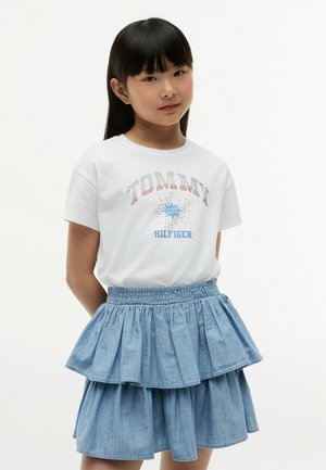 Ung pige med glat sort hår iført en hvid Tommy Hilfiger t-shirt og et blåt lagdelt denimskørt mod en ensfarvet baggrund.