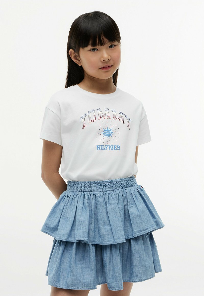 Jeune fille aux cheveux noirs et lisses portant un t-shirt blanc Tommy Hilfiger et une jupe en jean bleue à couches sur un fond uni.