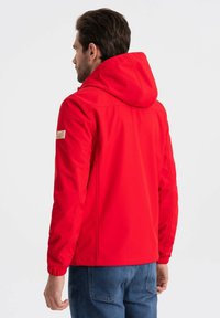 Rød jakke med hætte, der har en glat overflade, et lille side-label og et tætsiddende design. Båret over blå jeans.