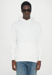 White hoodie featuring a front pocket and embossed "QUARTZ CO" logo. Model wears sunglasses and blue jeans, showcasing a casual look.