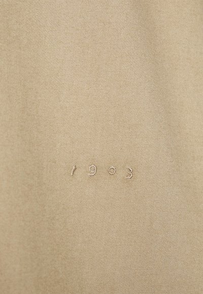 Tecido bege com uma textura suave e um brilho sutil, apresentando números bordados "1903" em um fio mais claro.