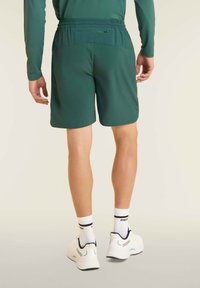 Pantalones cortos deportivos verdes con cinturilla elástica y bolsillos laterales, combinados con zapatillas blancas y calcetines con texto negro.