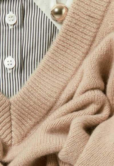 Cardigan en tricot rose doux, avec un col en V et une texture côtelée, associé à une chemise rayée en dessous et un accent de bouton en perles.