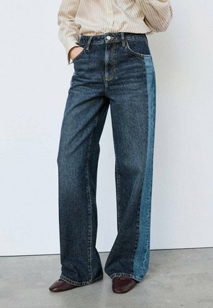 Persona que lleva vaqueros de pierna ancha bicolor con parte delantera oscura y paneles laterales más claros, combinados con zapatos marrones y una camisa de rayas.