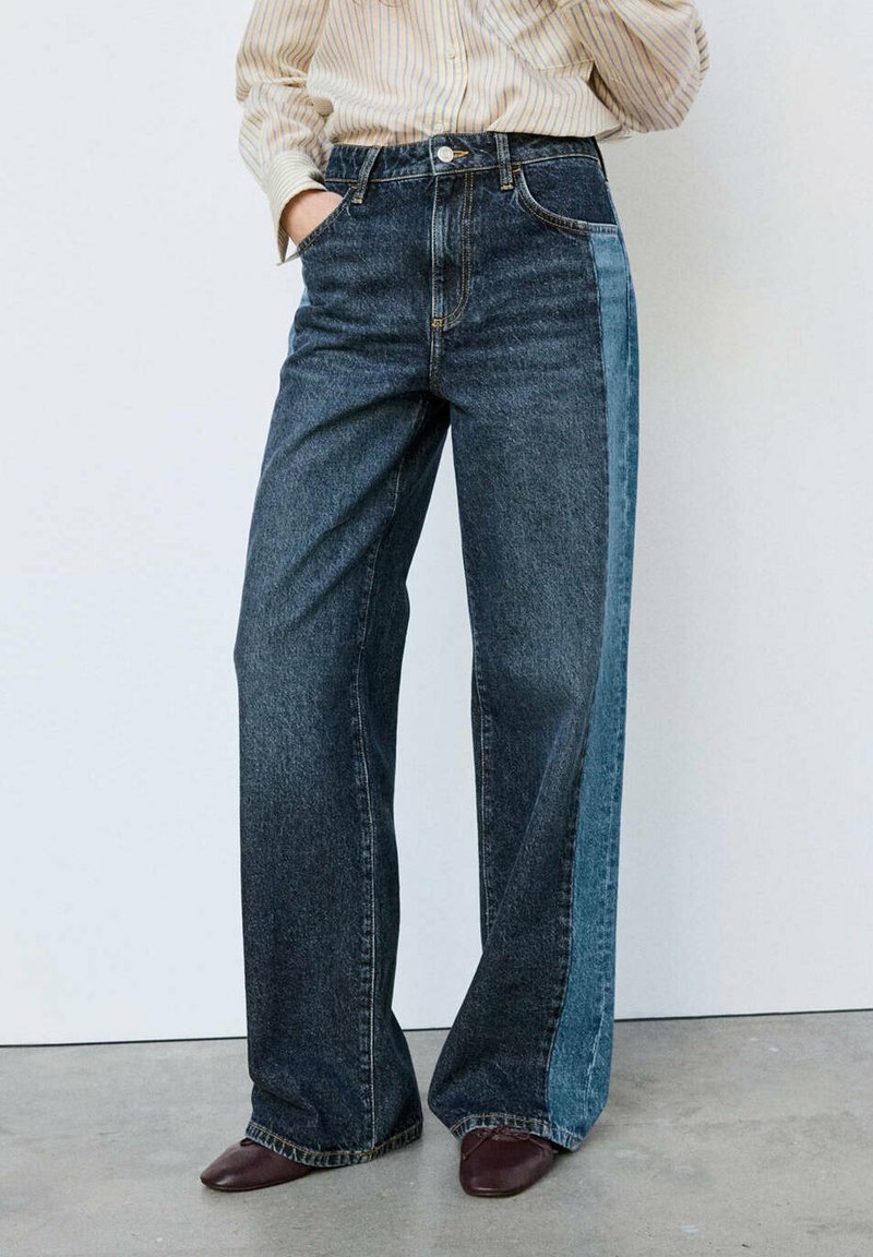 Personne portant un jean large bicolore avec un devant foncé et des panneaux latéraux plus clairs, associé à des chaussures marron et une chemise rayée.