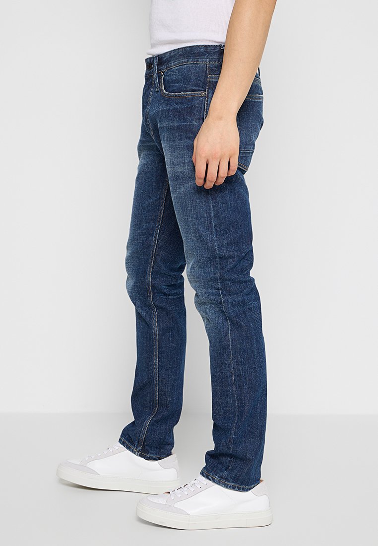 Donkerblauwe spijkerbroek met een slim fit, met een klassiek vijf-pocket ontwerp en subtiele stikseldetails. Gecombineerd met witte sneakers.