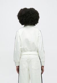 Fehér pulóver bordázott mandzsettákkal és derékpánttal, fekete oldalsó csíkokkal díszítve. Párban illik hozzá a hozzáillő fehér, magasított derekú nadrág.