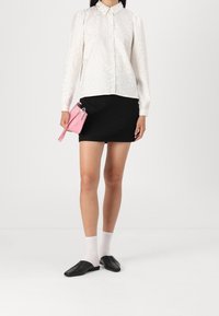 Chemise blanche texturée à manches longues avec col, associée à une jupe noire et des ballerines noires. Un petit sac à main rose est tenu dans la main.