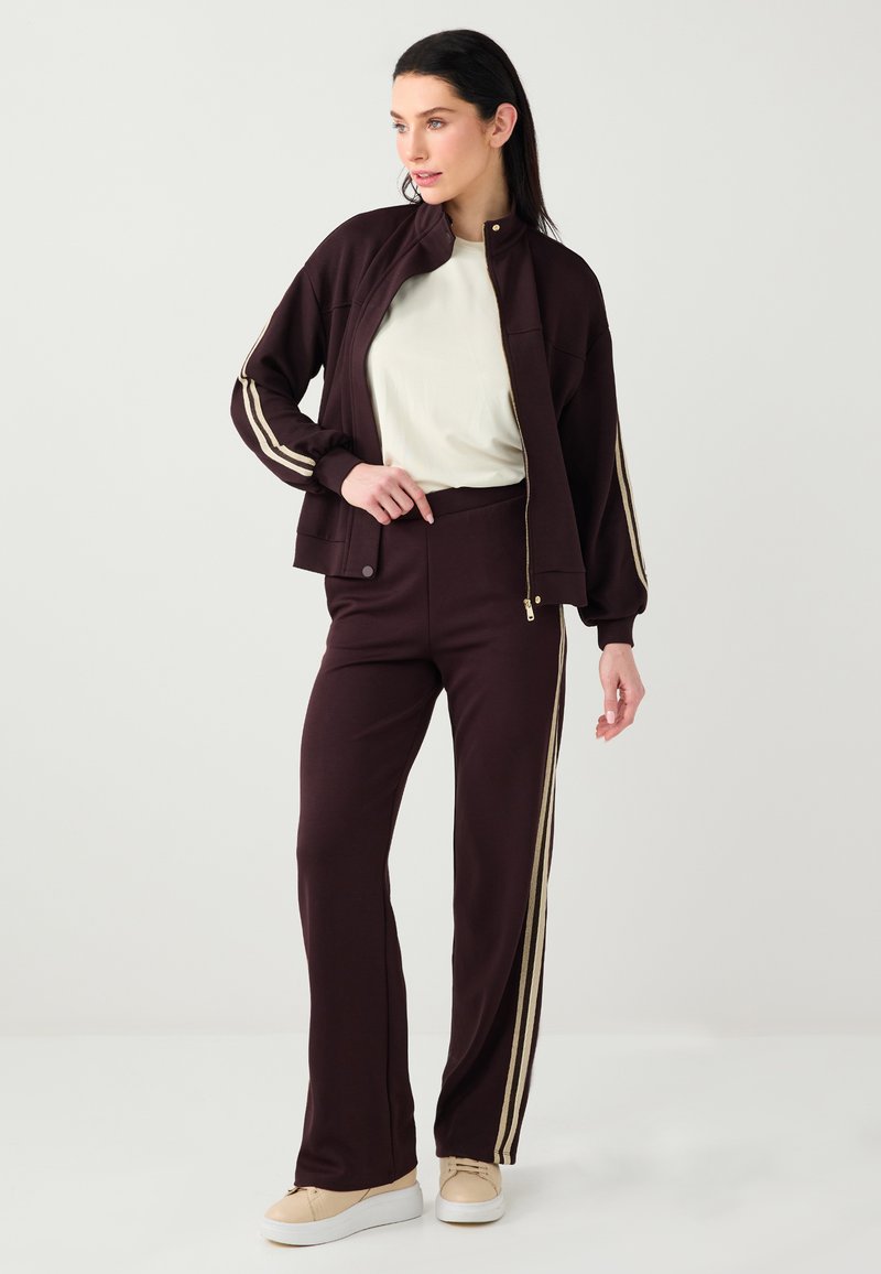 Survêtement bordeaux avec des rayures dorées, comprenant une veste zippée et un pantalon taille haute. Le tissu est doux avec une texture lisse.