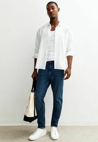 Camicia bianca a bottoni sopra una canotta bianca, jeans blu e sneaker bianche. Tiene una borsa a tracolla nera e crema. Sfondo semplice.