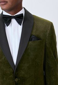 Mees, kes kannab tumerohelist sametist smokingut koos mustade satiinist litsakatega, valget voltitud dressipluusi, musta liblikat ja musta taskurätikut.