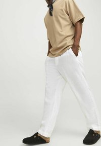 Mężczyzna w beżowej koszuli z krótkim rękawem, białych lnianych spodniach, czarnych butach typu slip-on i czarnym chustecie na szyi, stojący z rękami w kieszeniach.