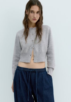 Fiatal nő hullámos barna hajjal szürke, gombos, rövidített kardigánt és laza, sötétkék, húzózsinóros nadrágot visel.