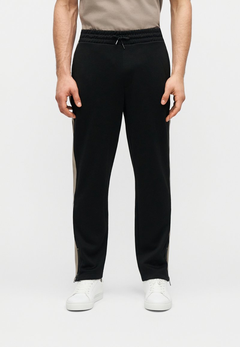 Άνδρας που φοράει μαύρο αθλητικό παντελόνι με μπεζ ρίγες στο πλάι και λευκά αθλητικά παπούτσια, στέκεται μπροστά από μια απλή φόντο.