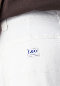 Witte werkbroek van duurzaam materiaal, met een zichtbaar blauw "Lee WORKWEAR" label op de achterzak en versterkte stiksels.
