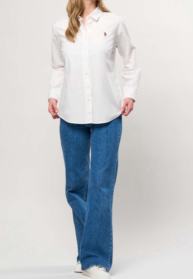 Femme portant une chemise blanche à manches longues boutonnée avec un petit logo brodé et un jean bleu à jambes larges avec des baskets blanches.