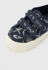 Sapatilha de lona azul-marinho com padrão de conchas do mar e estrelas-do-mar, apresentando duas tiras de fecho de velcro e uma sola de borracha branca texturizada.