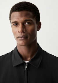 Chaqueta negra de cremallera con cuello, que presenta una textura mate y un diseño minimalista. El modelo tiene el pelo corto y rizado y una expresión neutral.
