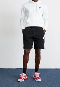 Homme portant un sweat à capuche blanc Nike, un short noir avec un petit cercle blanc, des chaussettes noires et des baskets Nike rouges et blanches, debout sur un tapis bleu.