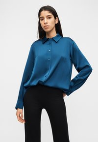 Γυναίκα στέκεται φορώντας μια σατέν πουκάμισο σε τιρκουάζ με κουμπιά και μαύρο παντελόνι, με το χέρι στη μέση και ουδέτερη έκφραση μπροστά σε απλό φόντο.