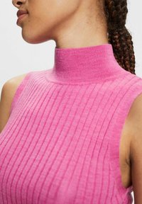 Lähikuva henkilöstä, joka on pukeutunut hihattomaan vaaleanpunaiseen, ribattu korkean kauluksen villapaitaan, ja hänen letitetyt hiuksensa on vedetty taakse.