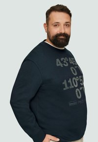 Sweatshirt bleu marine avec un col rond, présentant un grand texte gris et des coordonnées géographiques. Texture lisse et coupe décontractée.