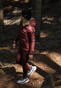 Giubbotto puffer color bordeaux con logo, leggings bordeaux abbinati e scarpe da ginnastica grigie e bianche. Situato in un'area boschiva con aghi di pino.
